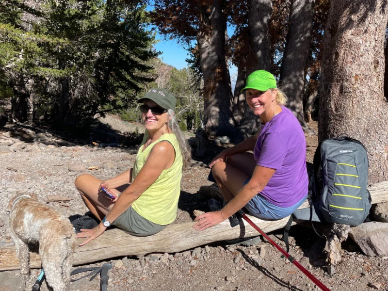 Two women hikers and a dog sit on a log resting during the Be Warrior Challenge Hike in 2023